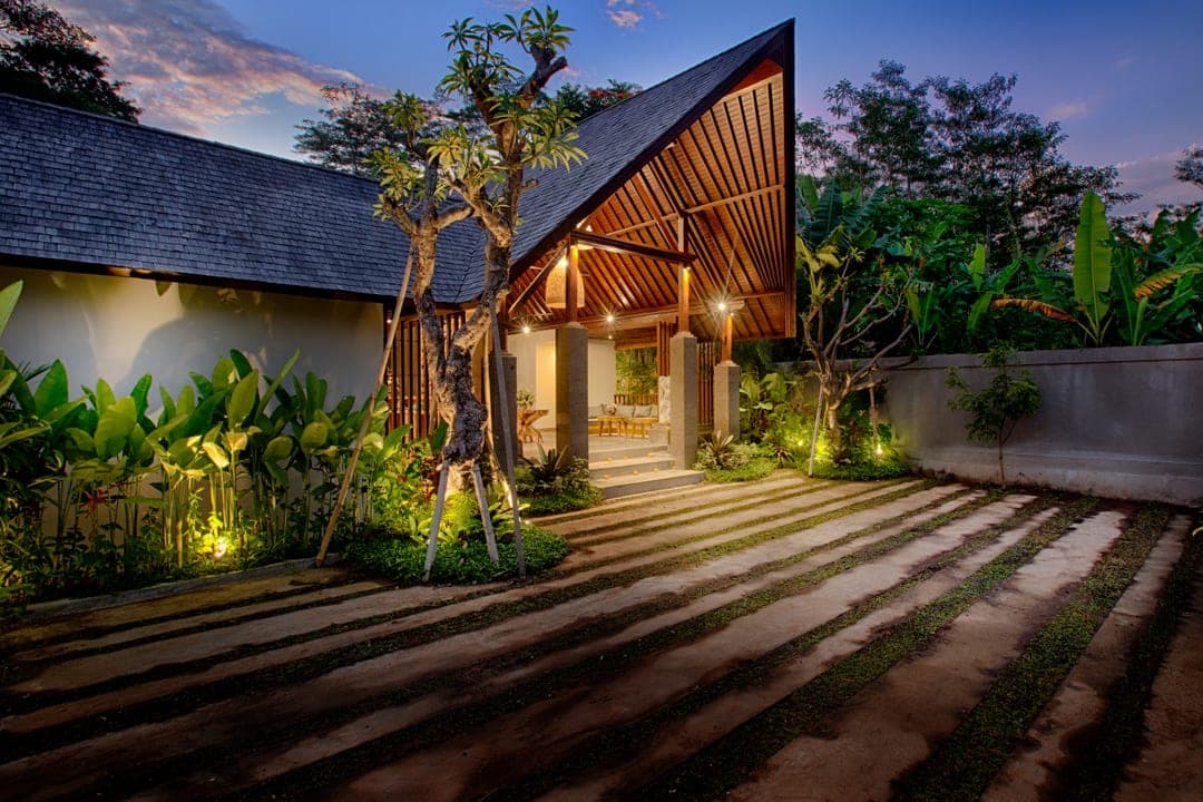Villa entrance at dusk: a steep timber A-frame pavilion lit from within, a frangipani tree and a stone path set into strips of grass