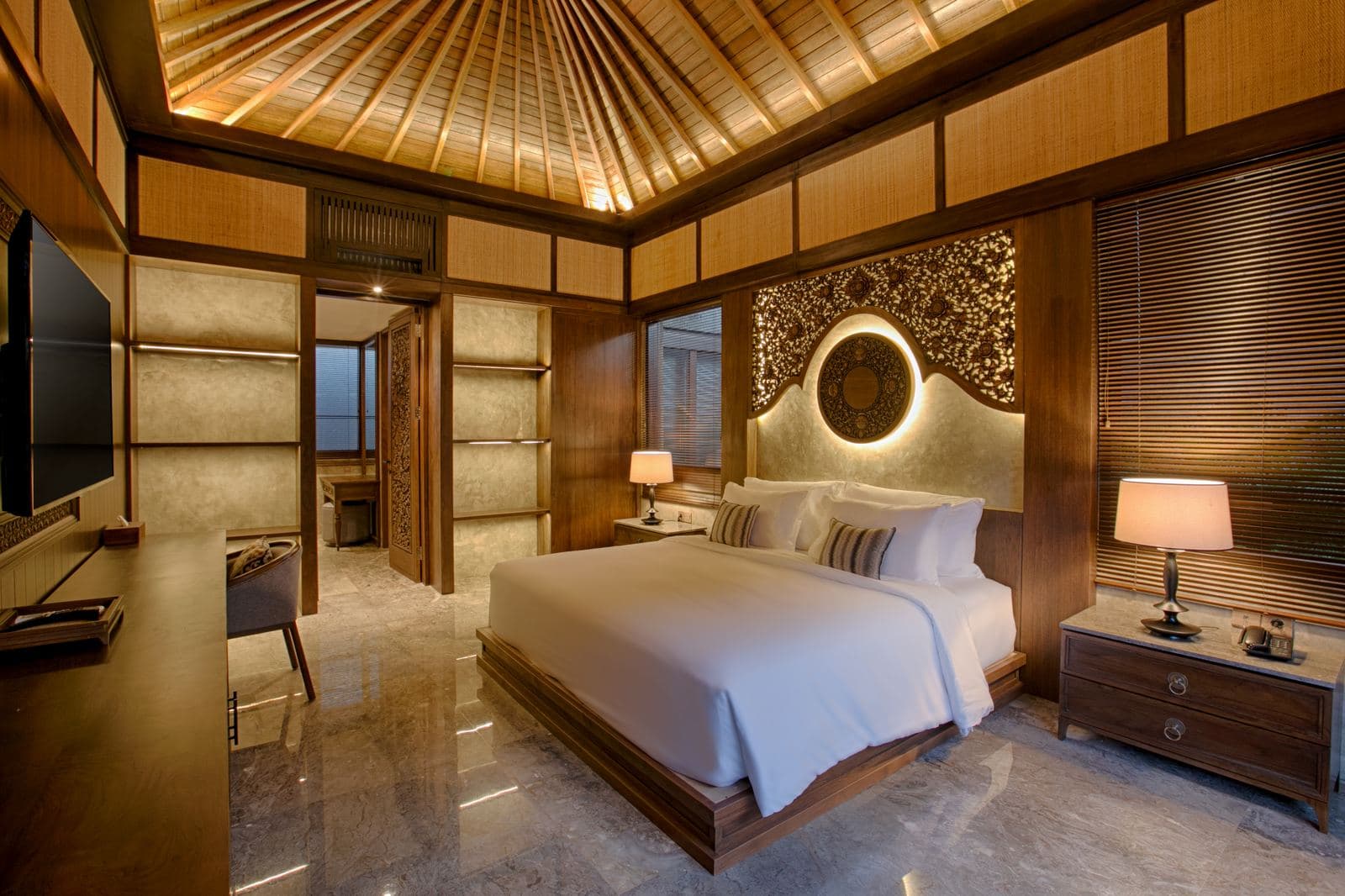 Suite bedroom under a high thatched ceiling, a carved and gilded headboard lit from behind, timber screens and matching bedside lamps