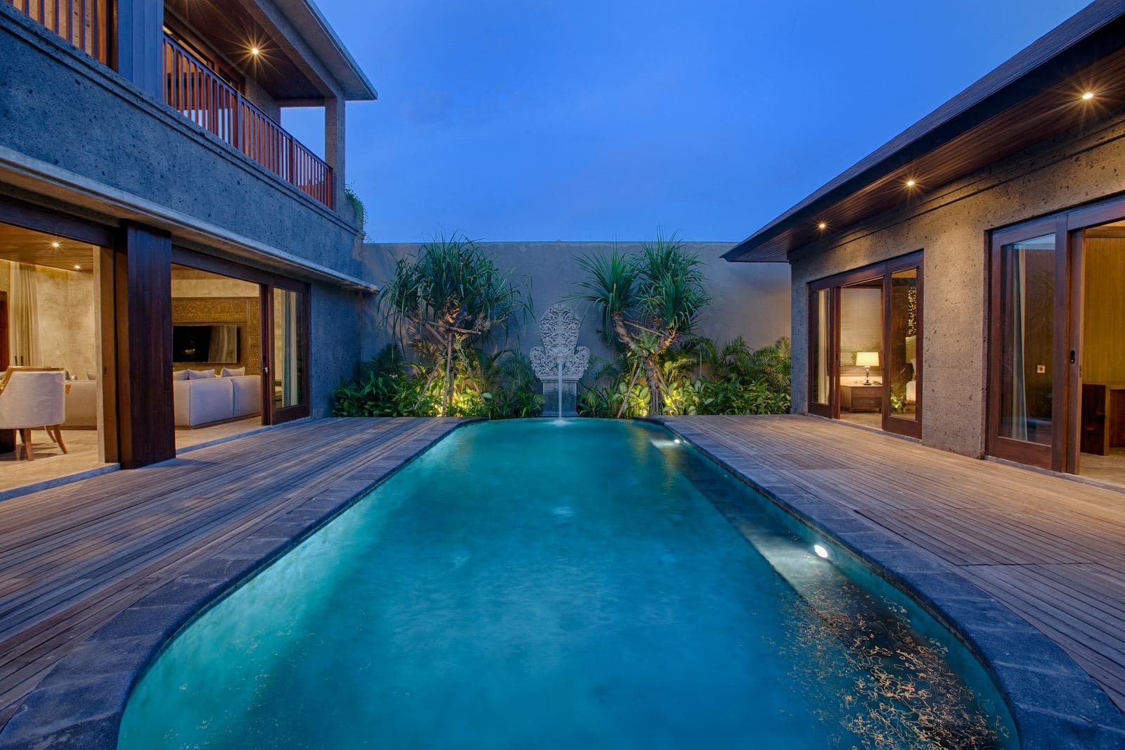 Long pool at dusk between two-storey villa wings, a water spout at the far end and deck lights doubled in the water