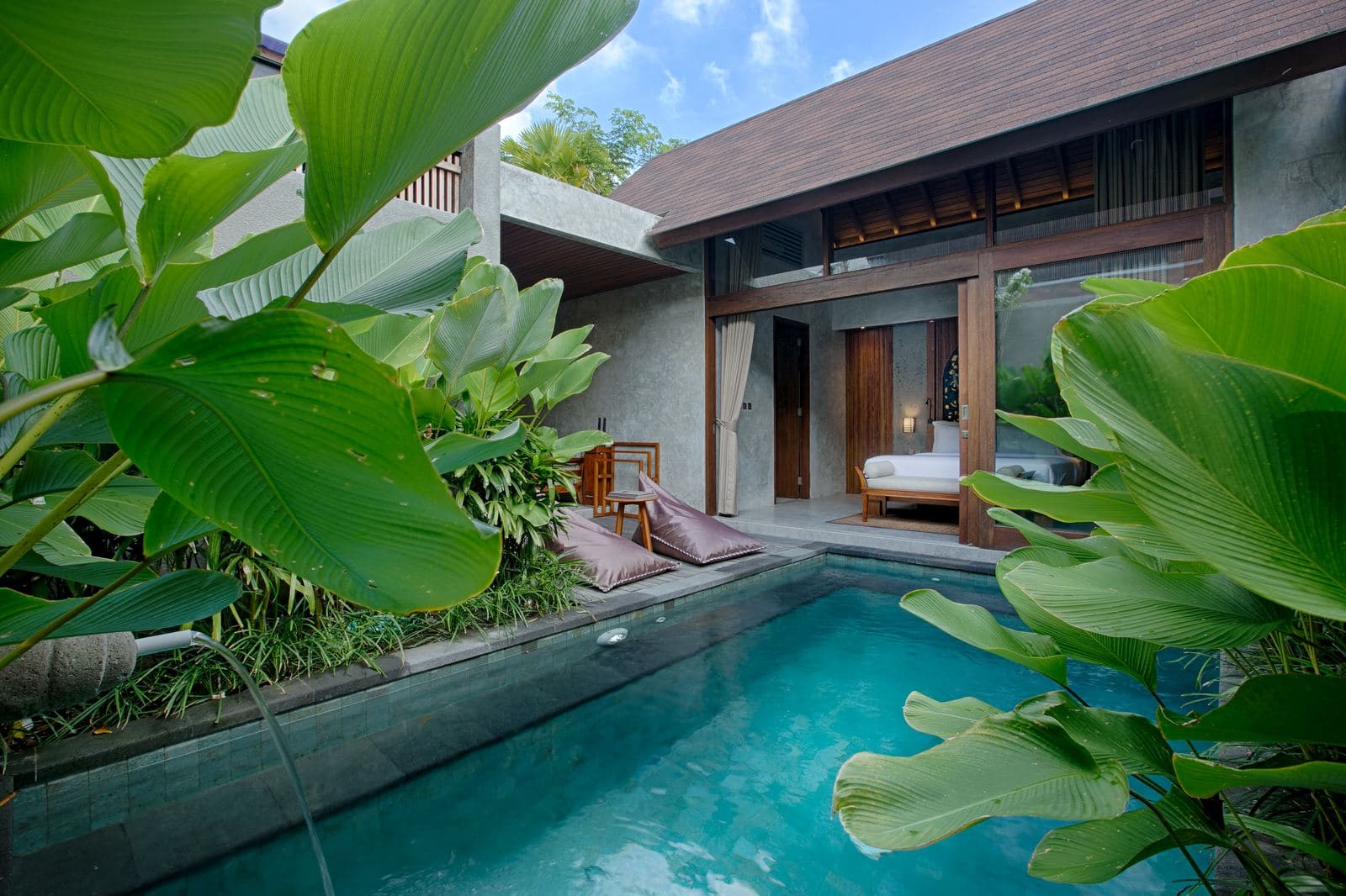 Private pool seen past oversized green leaves, the bedroom pavilion open behind it under a thatched roof