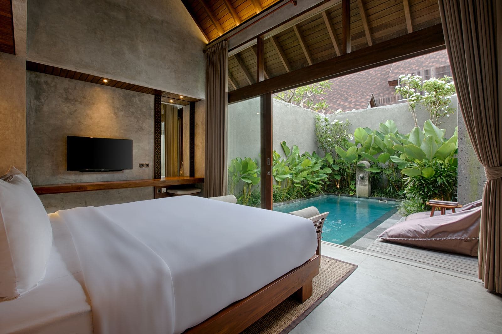 Bedroom at dusk with a white bed facing open glass doors onto its private pool, a television mounted on the stone wall
