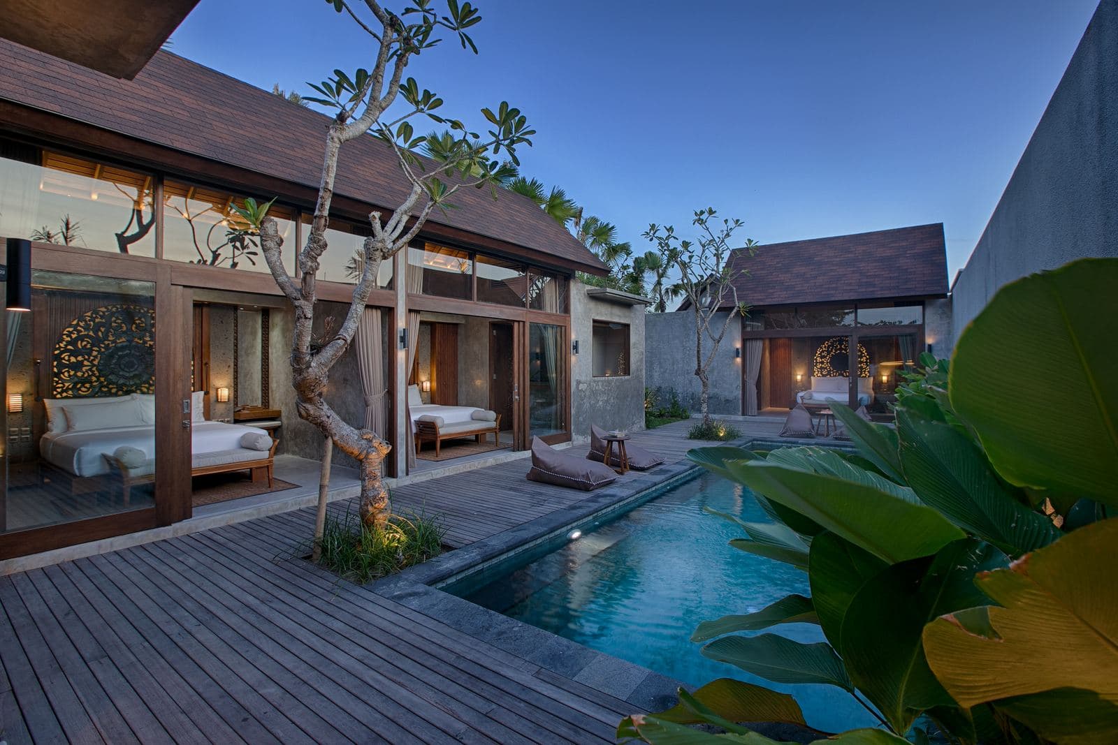Three-bedroom villa courtyard in blue evening light, a frangipani tree over the deck and lit bedrooms along two sides of the pool