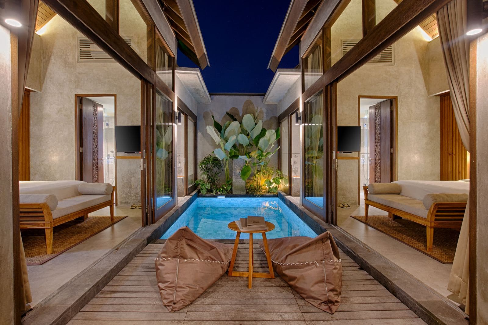 Villa courtyard at dusk: a lit pool running between two open pavilions, beanbags and a round timber stool on the deck beside it