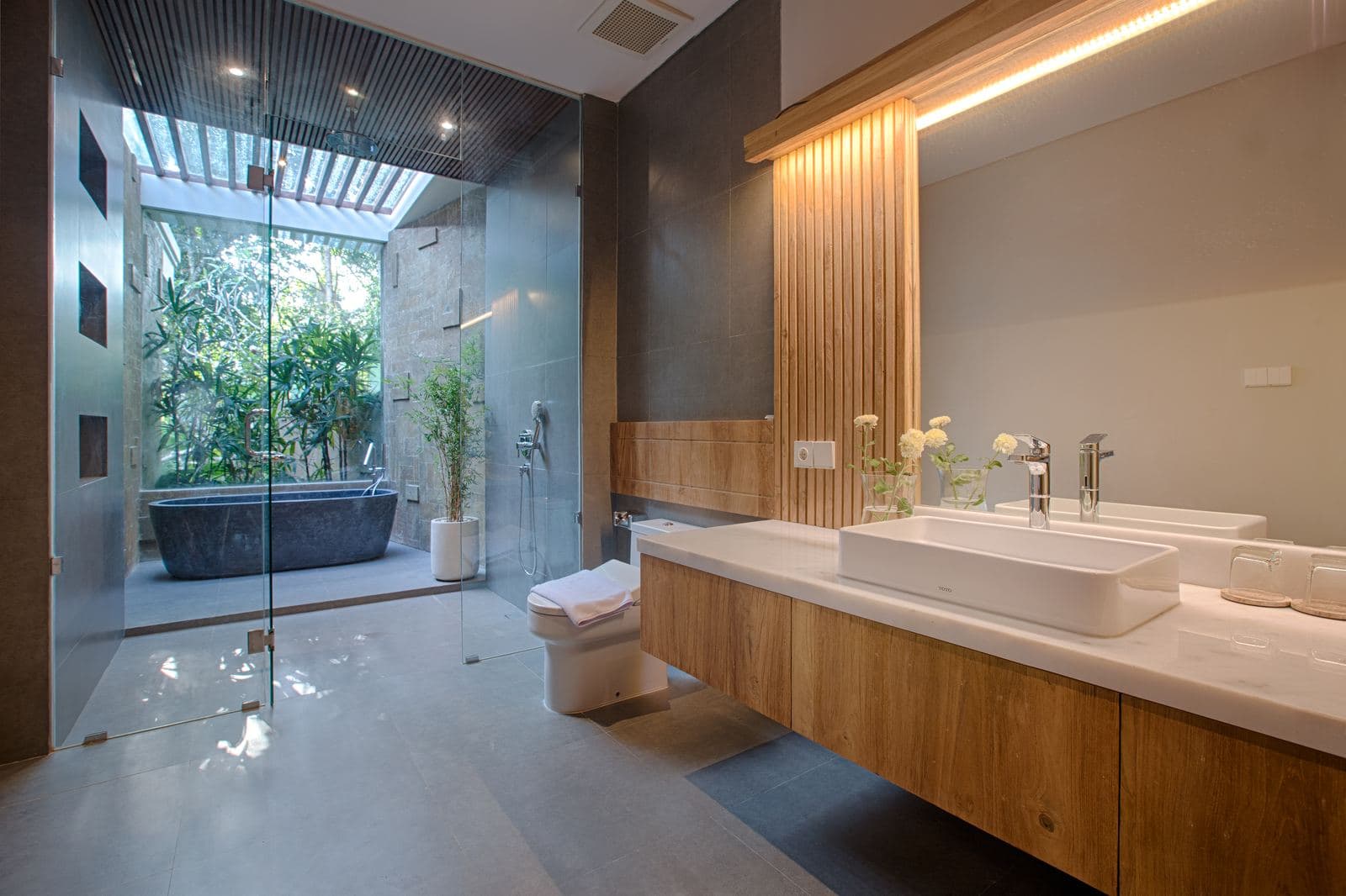Bathroom with a long timber vanity and twin basins under a backlit mirror, and a stone tub behind glass in a planted outdoor shower