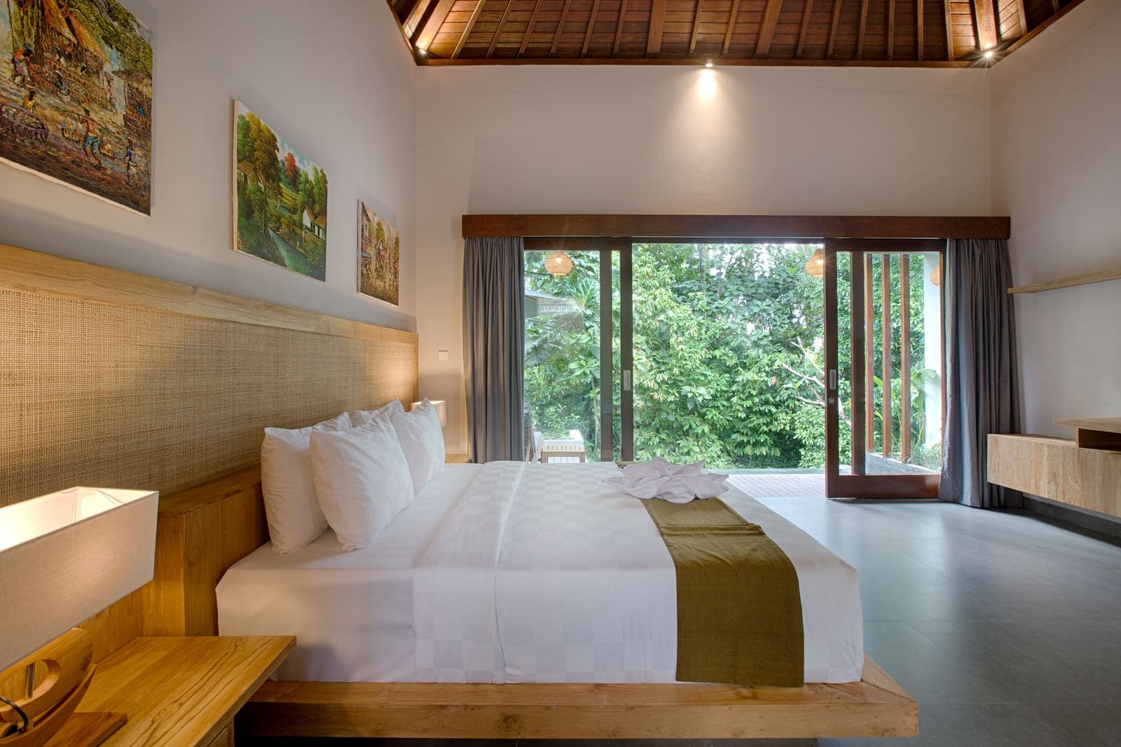 Bedroom with a platform bed against a woven headboard wall, paintings above it and sliding doors onto a green garden