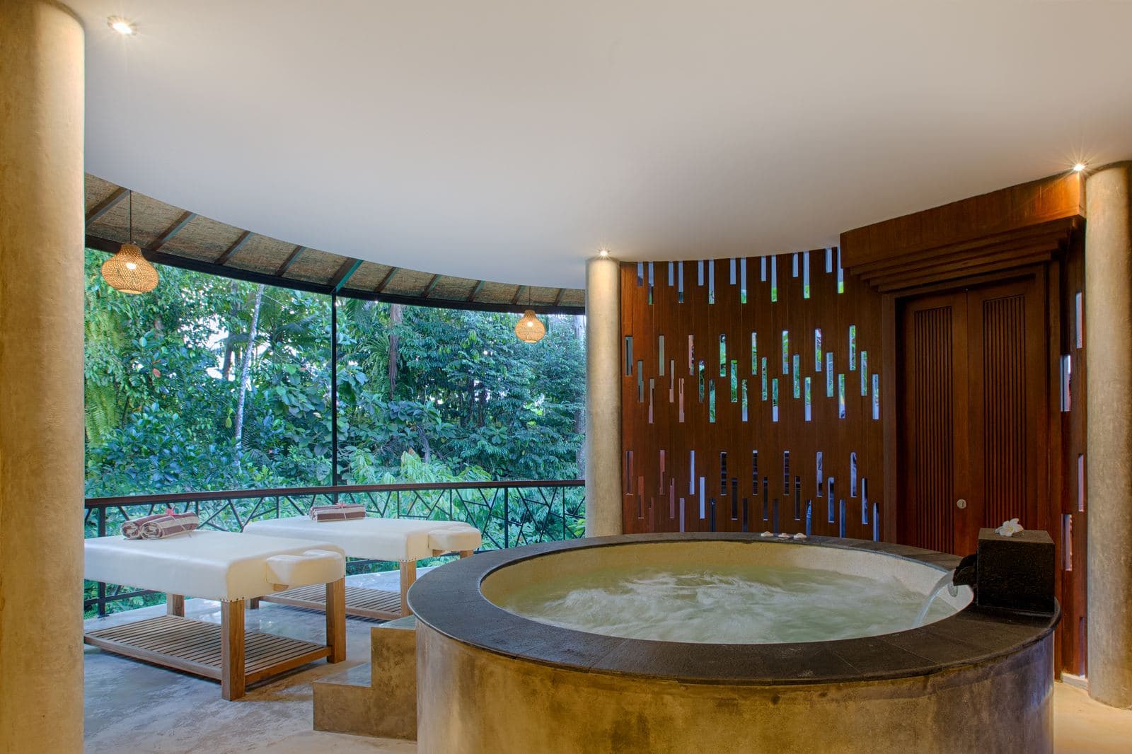 Round stone jacuzzi filled to the brim beside two massage beds, open to the jungle over a curved balcony rail