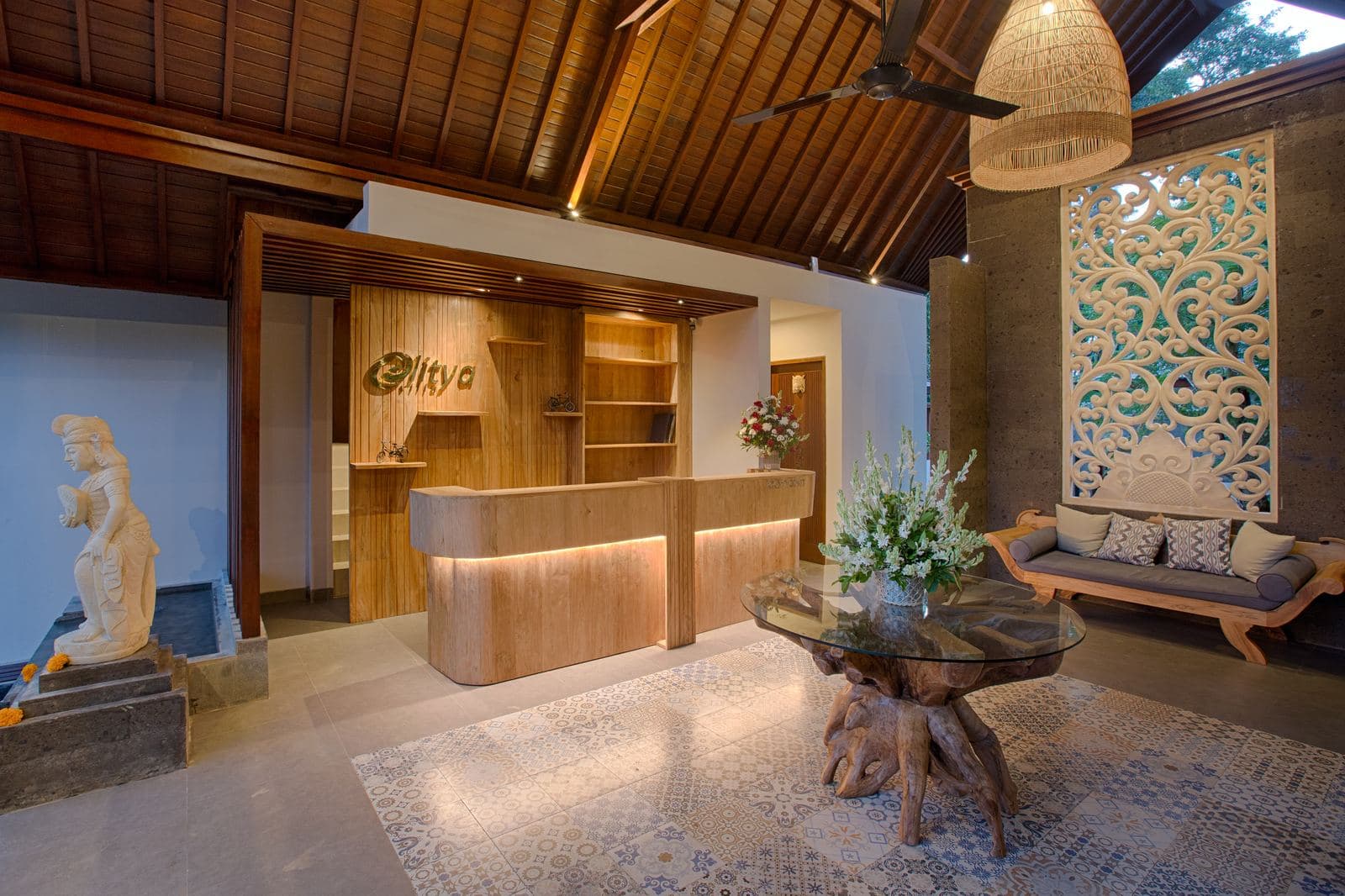 Lobby with a curved timber reception desk, a carved stone guardian statue, a glass-topped root table on a patterned rug and a daybed under a carved panel