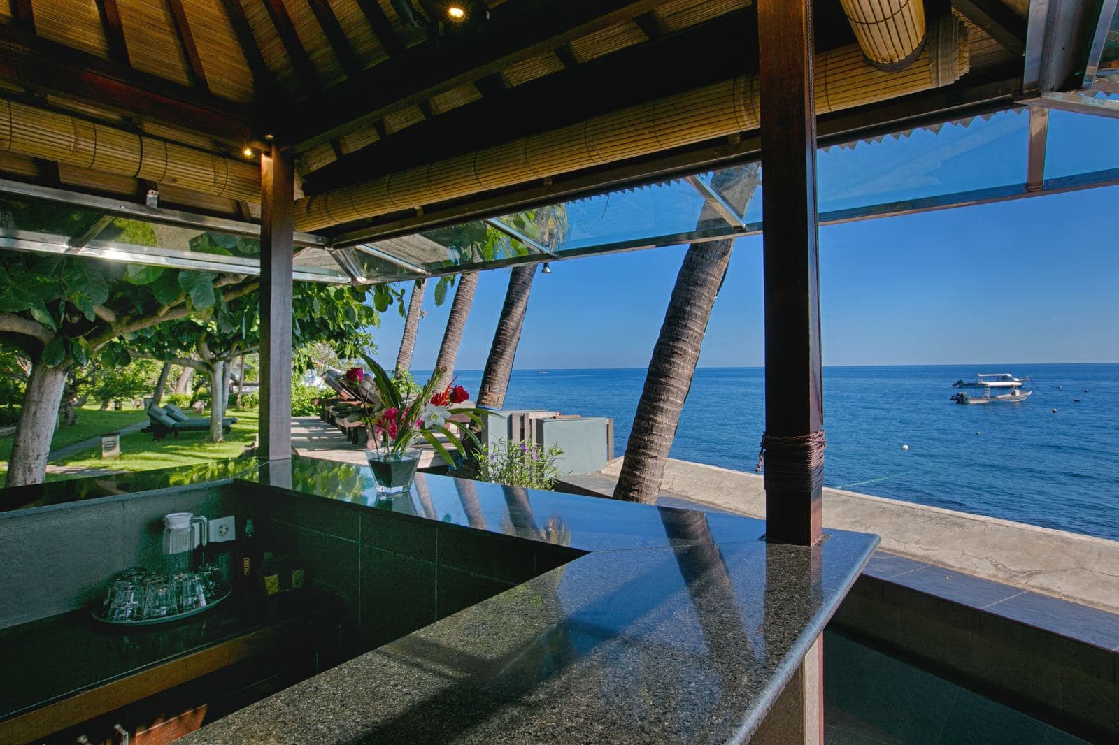 Open-sided bar with a polished stone counter looking straight out to a calm sea, palms framing the view and a fishing boat moored offshore