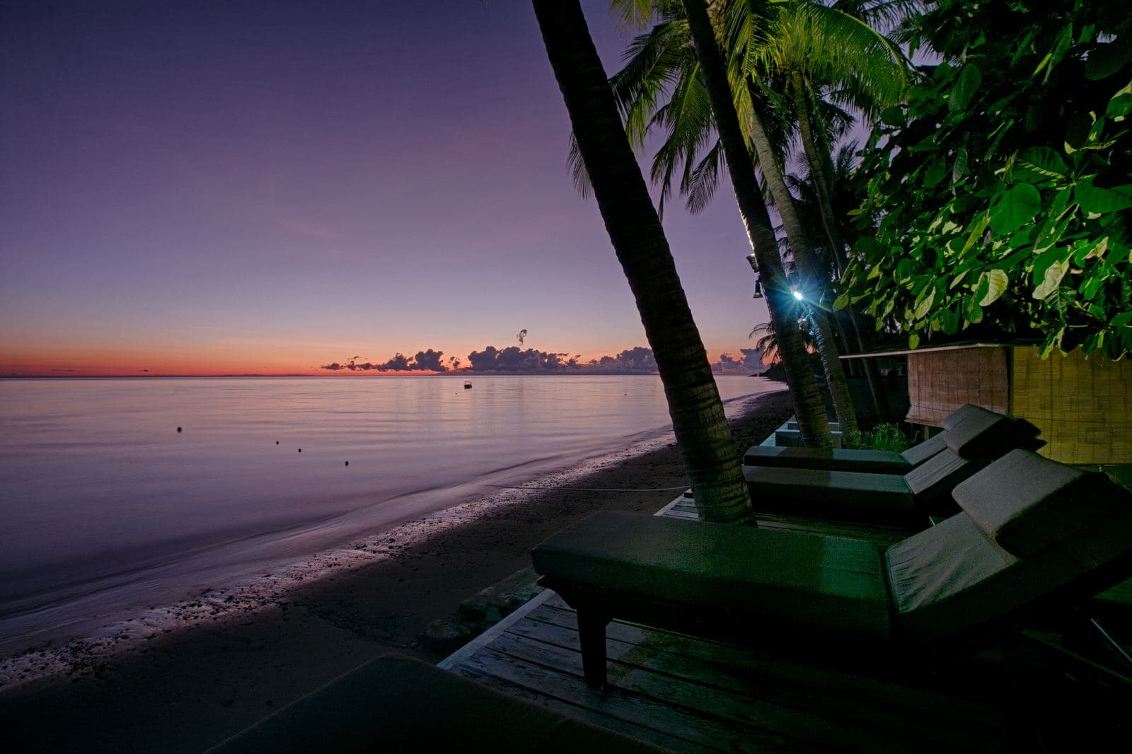 Dark sand beach at sunrise, the sea flat and pale to the horizon, two loungers on a timber deck beneath a palm
