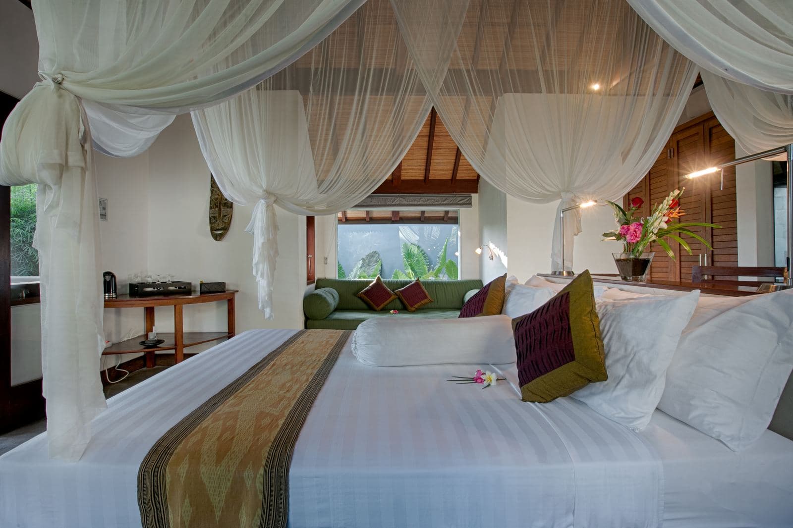 Bungalow bedroom with a four-poster bed under white mosquito netting, gold and plum cushions, and a sofa at the foot of the bed