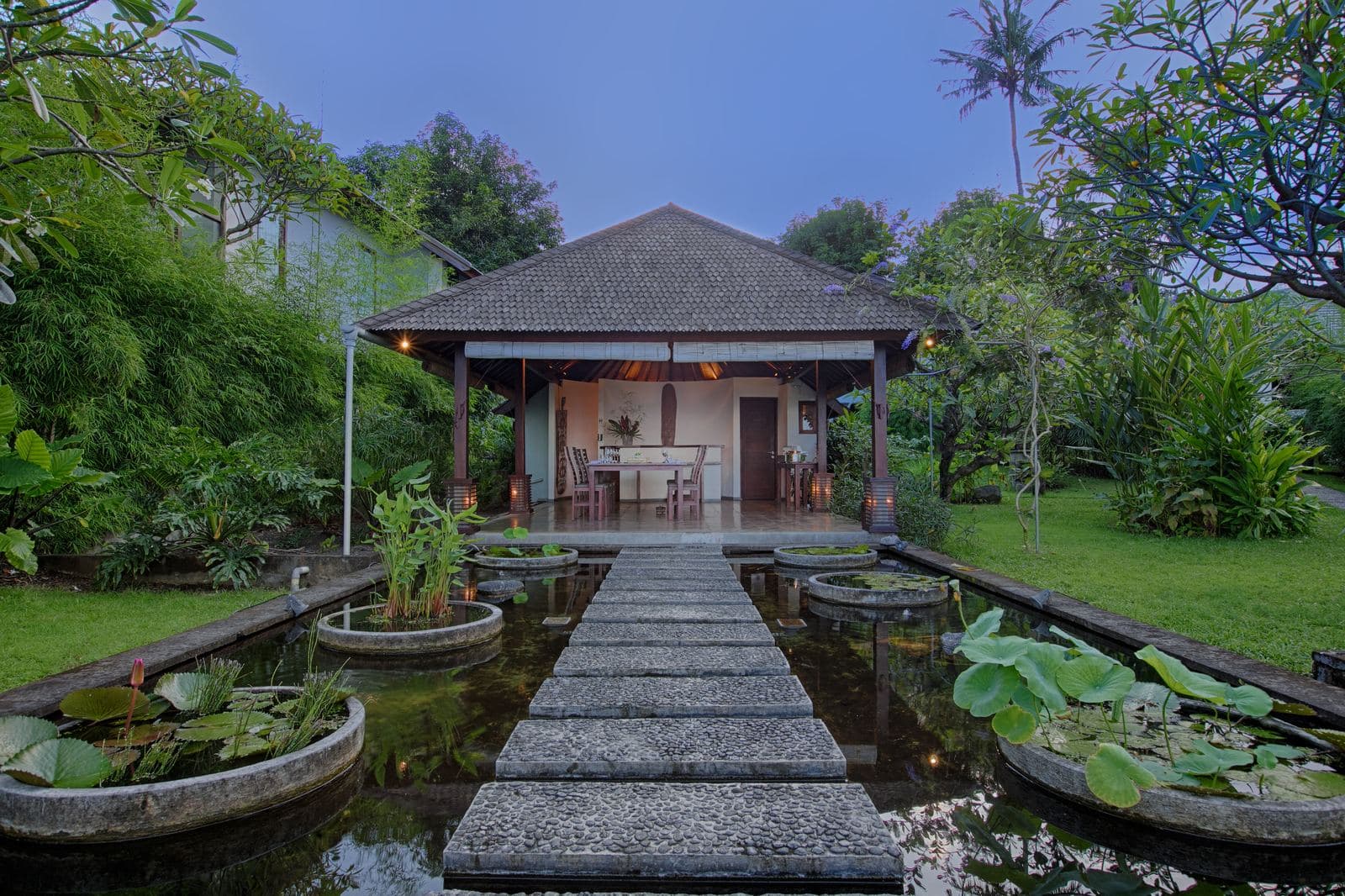 Stone path across a lotus pond leading to an open restaurant pavilion with a shingled roof, garden either side in evening light
