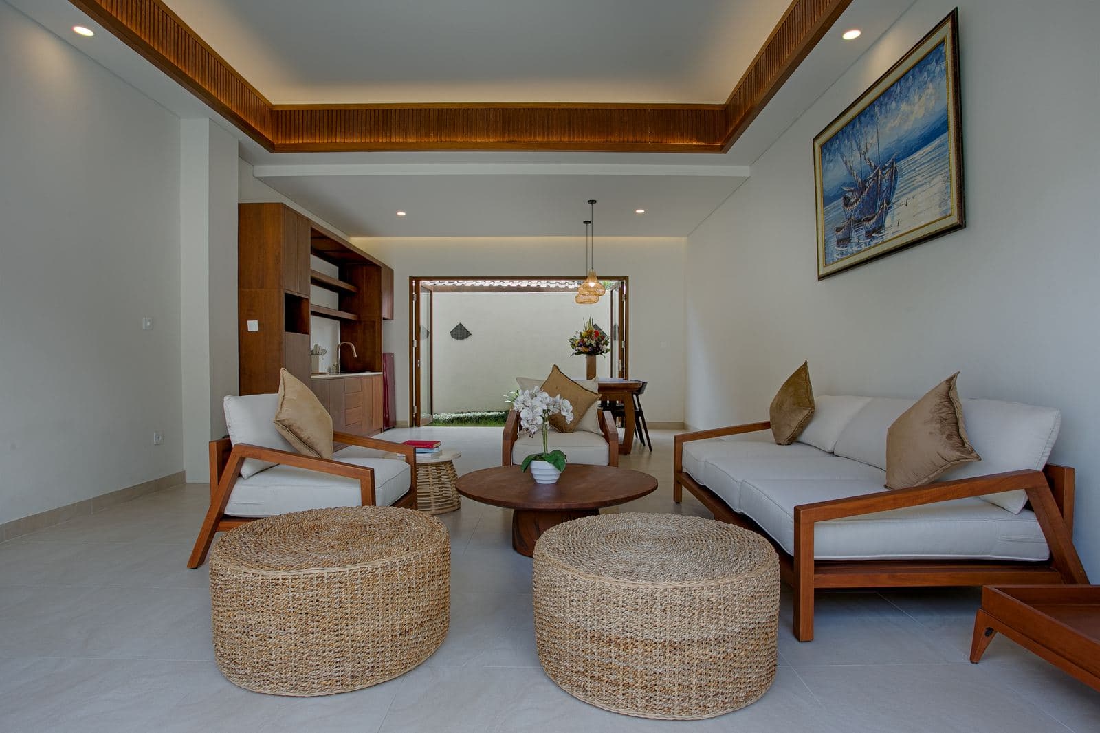 Living room with two timber-framed sofas facing a round coffee table, woven rattan ottomans, and a dining table beyond the open doors