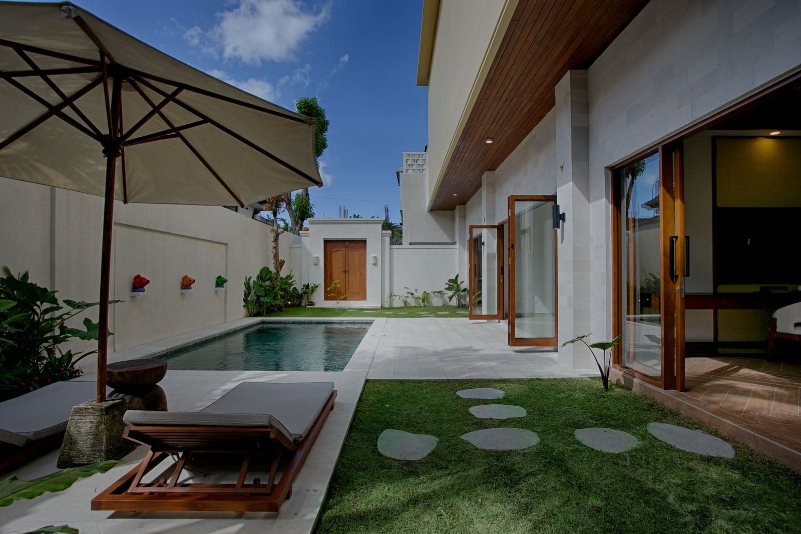 Side garden with a lap pool, a lounger under a white parasol and stepping stones across the lawn, the villa's glass doors folded open along it