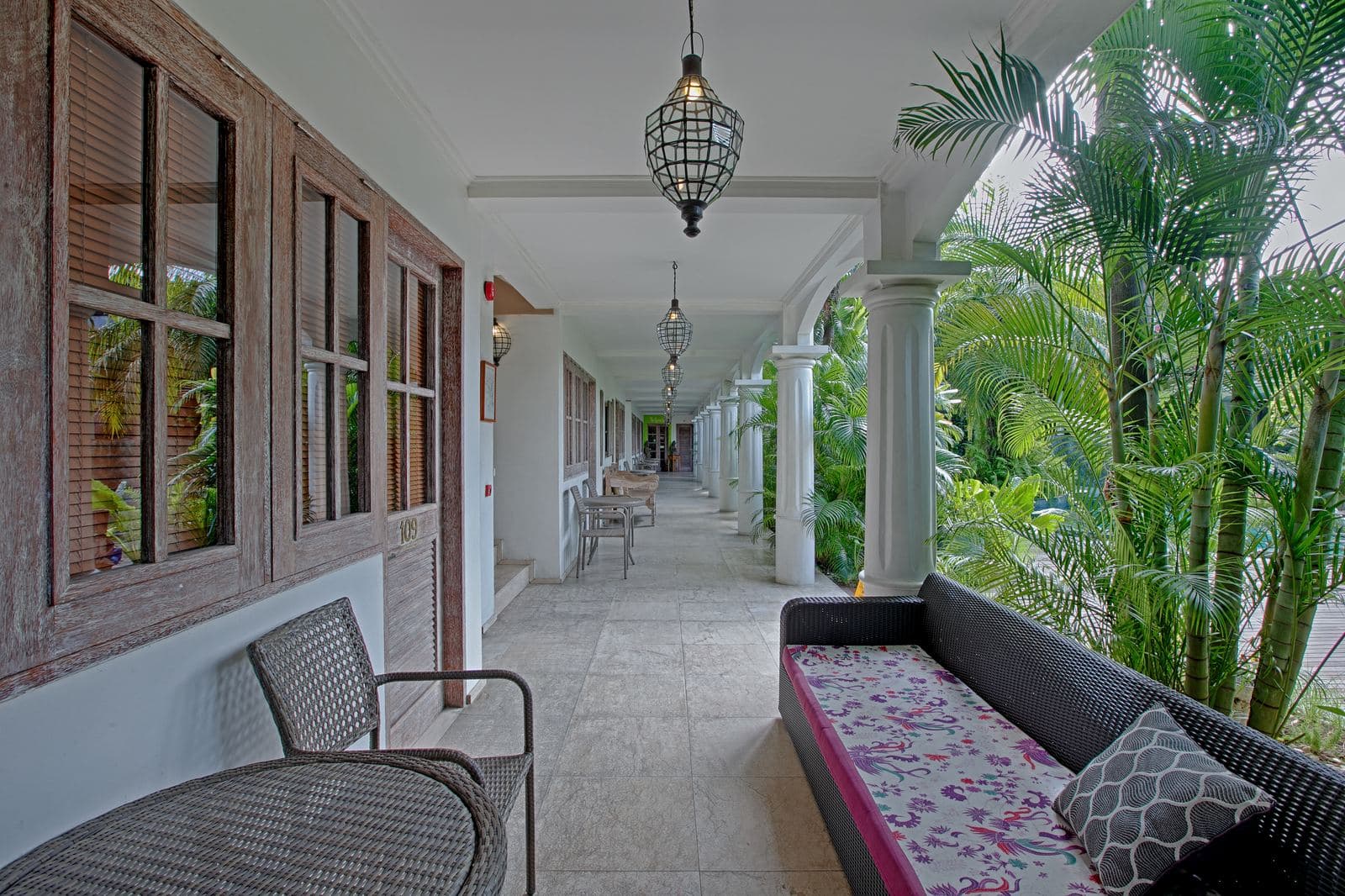 Ground-floor colonnade with lantern pendants, rattan armchairs and a long cushioned bench, palms crowding the open side
