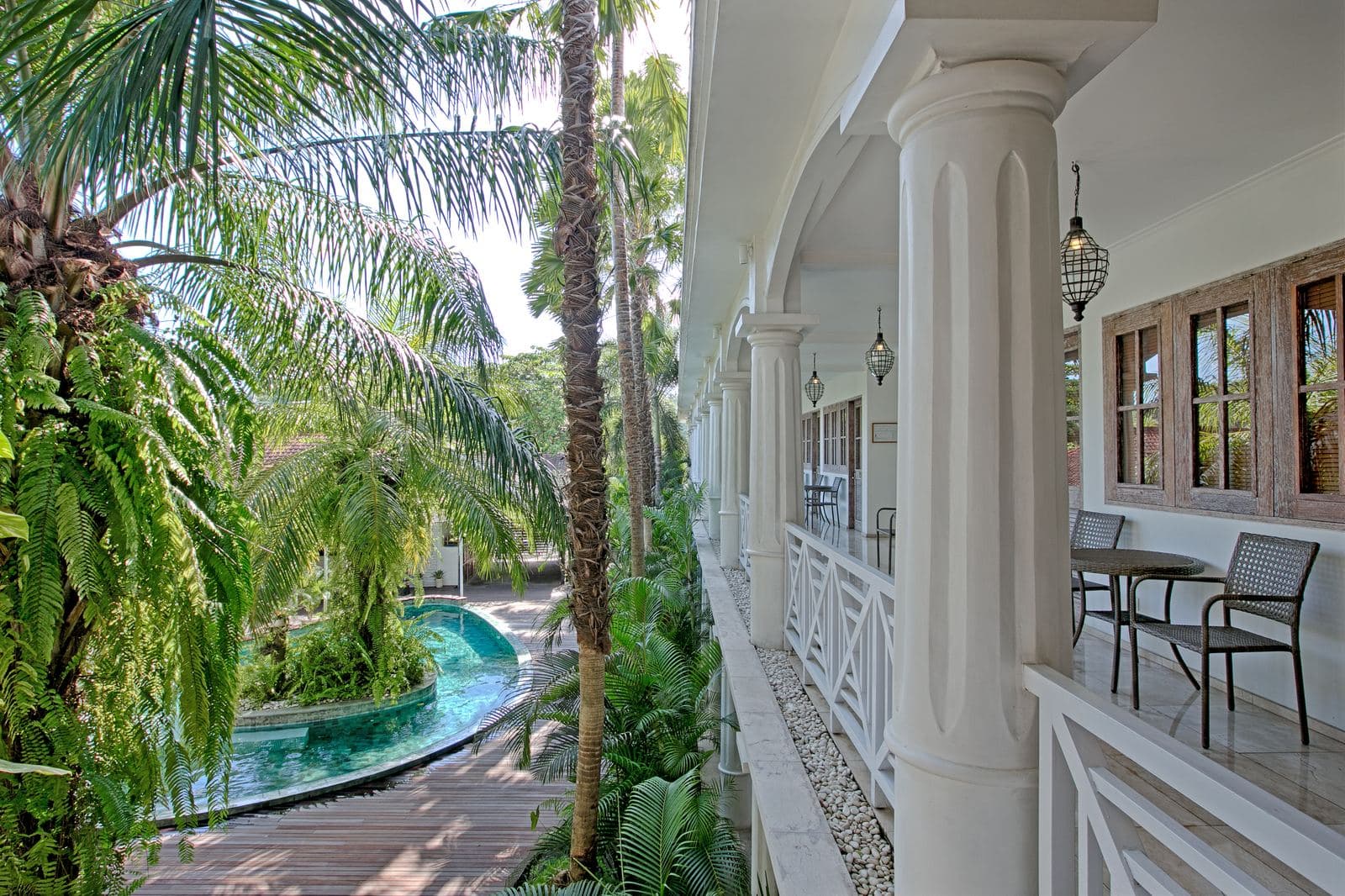 Upper-floor colonnade with white columns and a timber balustrade, looking down through palms to the curved pool below
