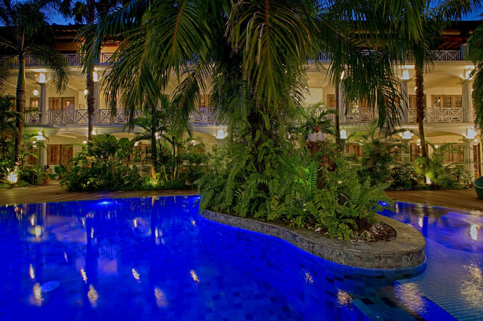 The same pool at night, lit deep blue around a planted island of palms, the guest wings glowing warm behind it