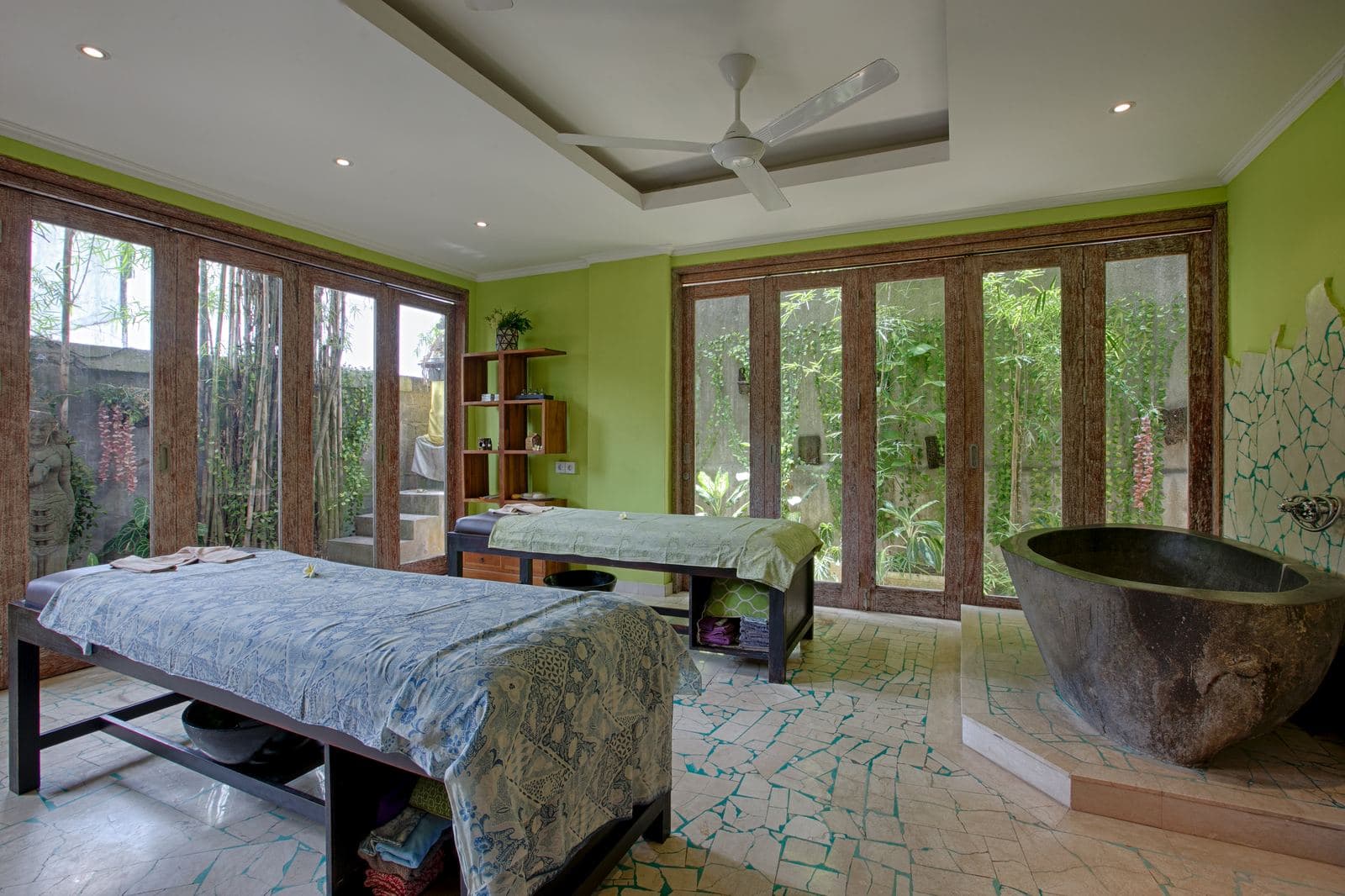 Spa room with two massage tables, a carved stone bath, green walls and glass doors on two sides opening onto the garden