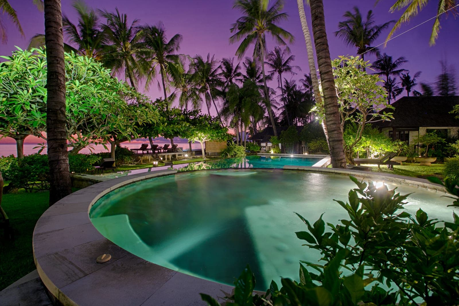 Beachfront villa pool at dusk under coconut palms, the water lit green against a deep purple sky