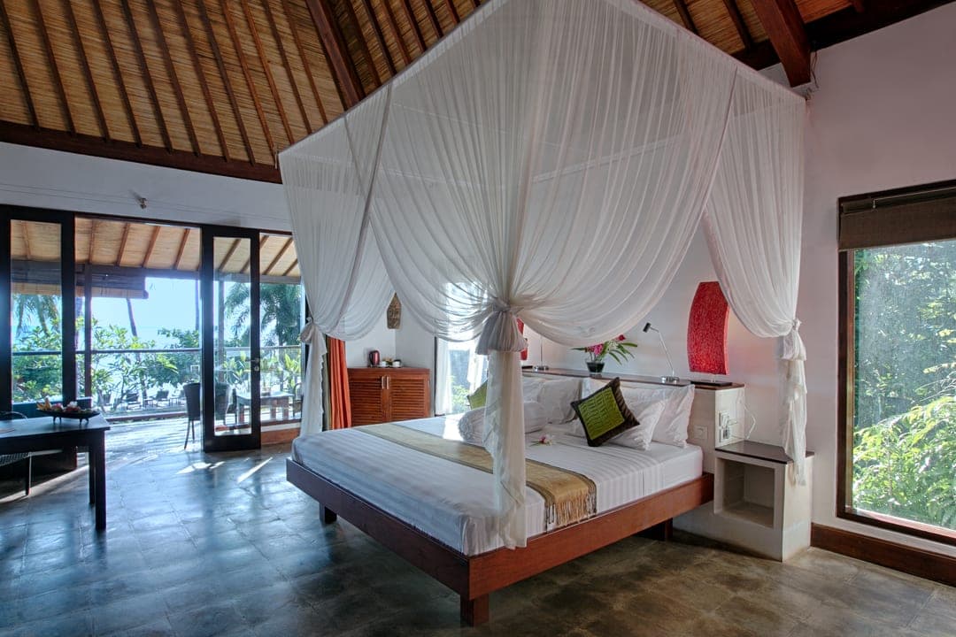 Villa bedroom with a four-poster bed under white netting and a high thatched ceiling, opening onto a terrace and the sea