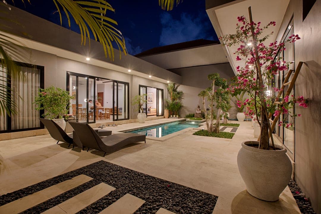 Courtyard villa at night with a lap pool, loungers and flowering bougainvillea, the living and dining rooms lit behind glass doors