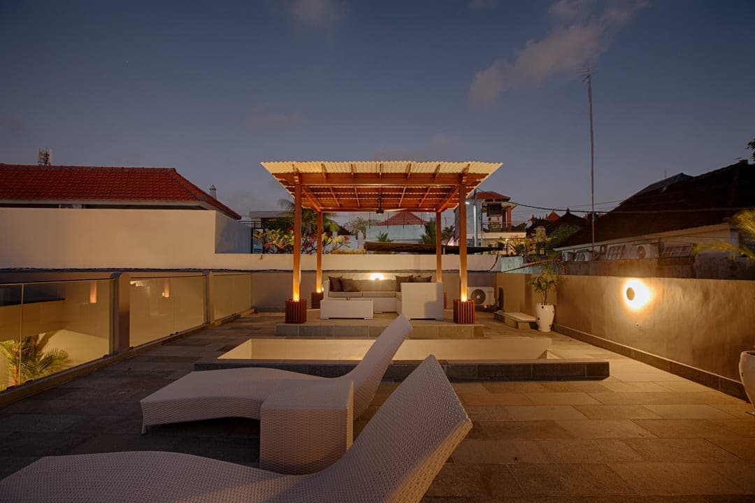Rooftop terrace at dusk with a timber pergola, white seating and a plunge pool, rooftops and palms beyond