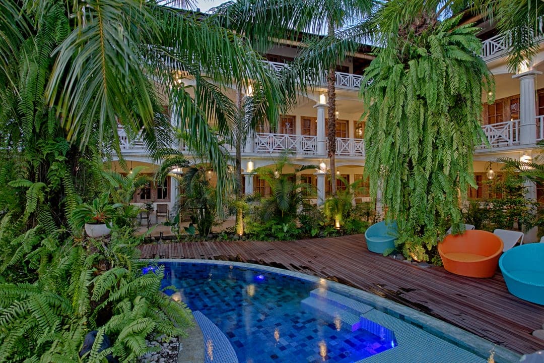 Two-storey colonial resort courtyard seen through ferns and palms, with a curved pool, timber deck and coloured tub chairs