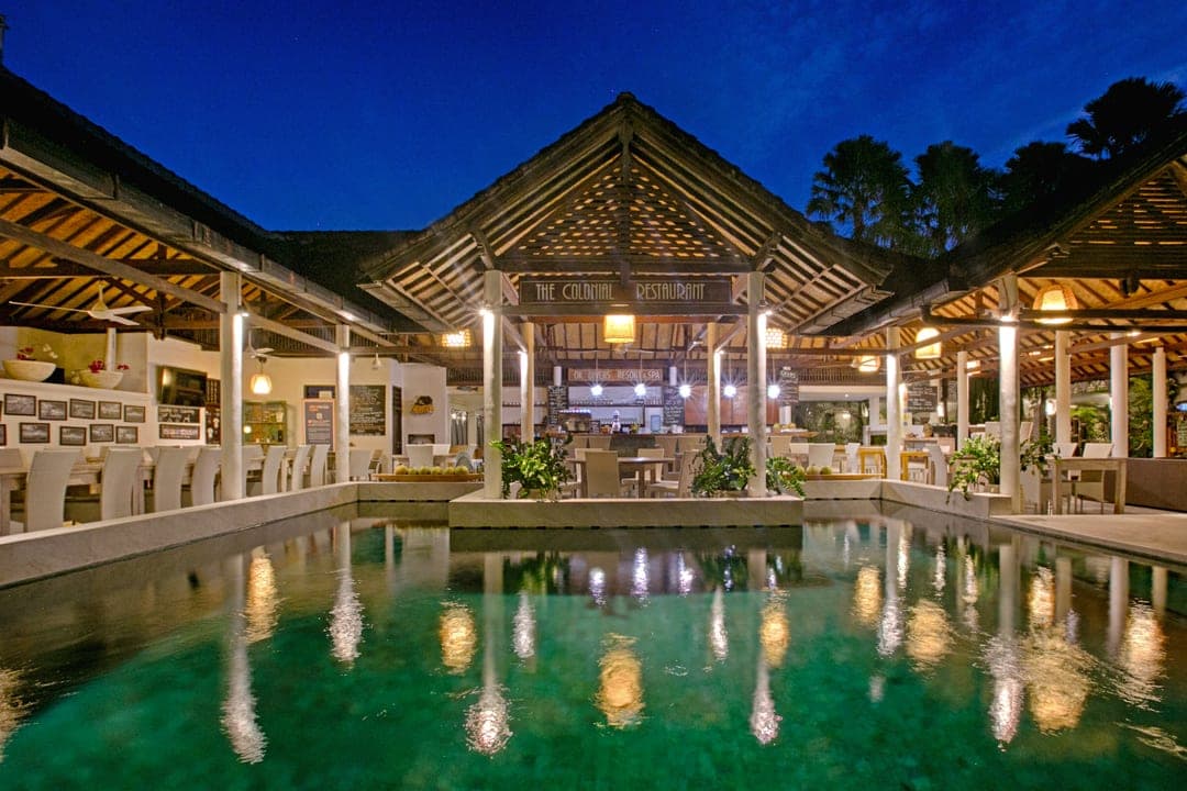 Open-sided resort restaurant at dusk, its lit timber roof and columns reflected in the pool in front of it